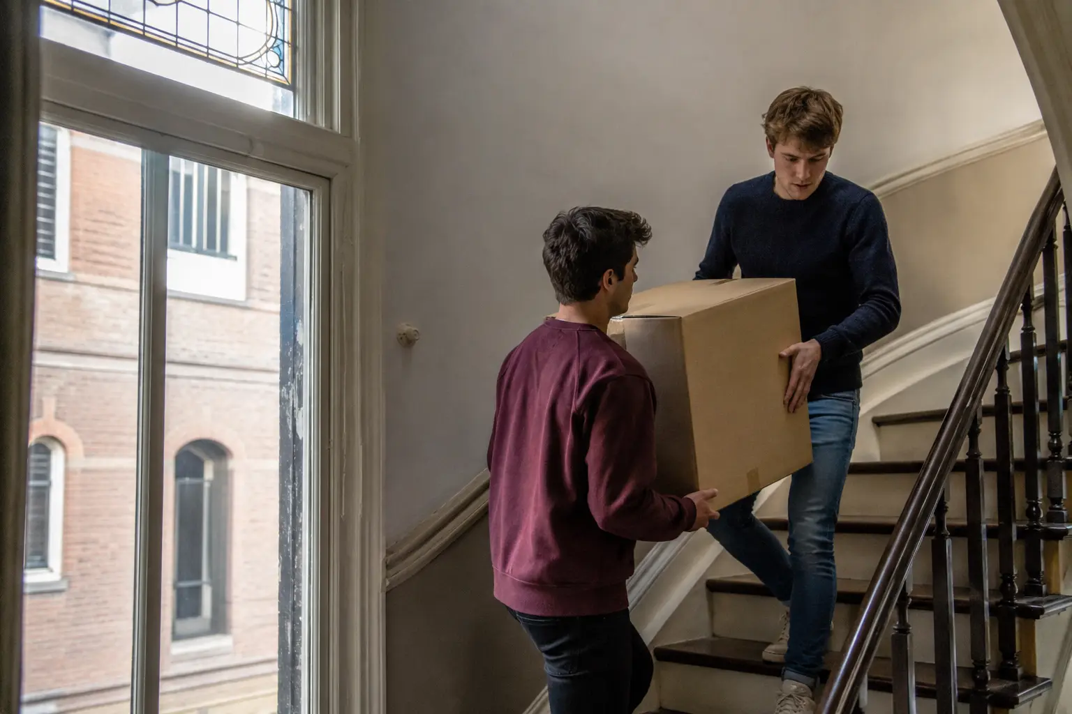 Twee jonge verhuizers dragen samen een doos door een Nederlands trappenhuis